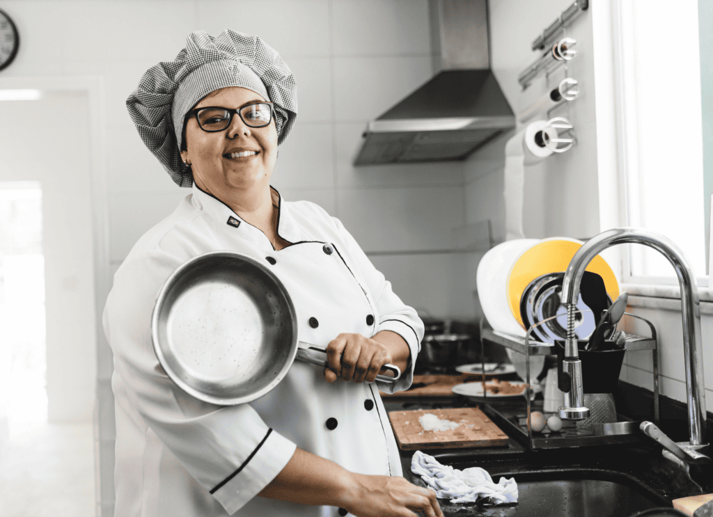 A cheerful chef wearing glasses and a grey checkered hat smiles while holding a stainless steel pan in a bright kitchen. She wears a white chef’s coat with black buttons. Behind her, a tidy kitchen workstation includes a dish rack with yellow and white dishes, utensils in a container by the sink, and ingredients on the counter. Natural light streams in from a window, highlighting the cleanliness and joy of the cooking environment.