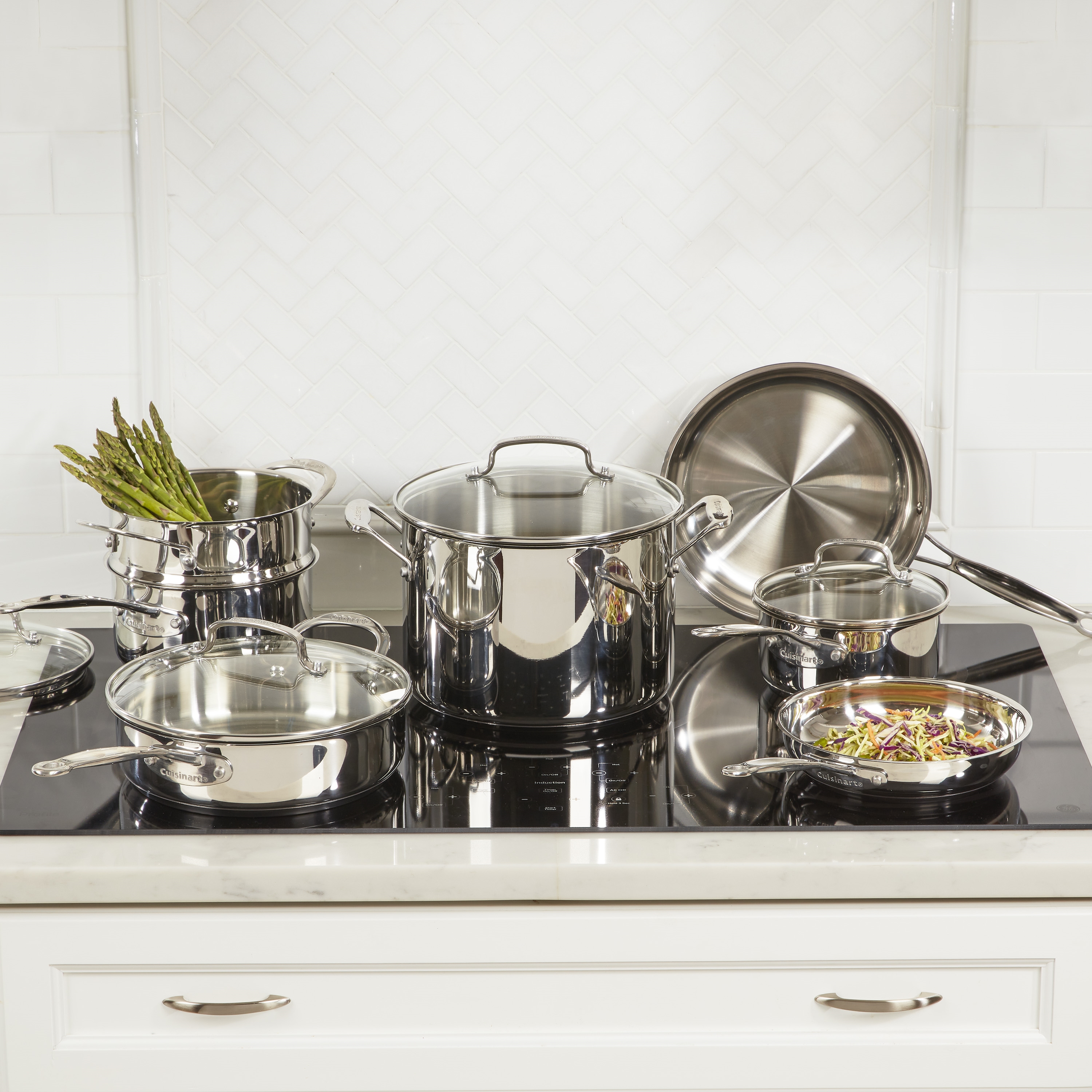 A Cuisinart stainless steel cookware set displayed on a marble countertop against a white herringbone tile backsplash. The set includes various pots and pans with reflective surfaces and lids. One pot holds fresh asparagus, and a small pan contains colorful vegetable stir-fry, highlighting the cookware's functionality and elegant design in a stylish kitchen setting.