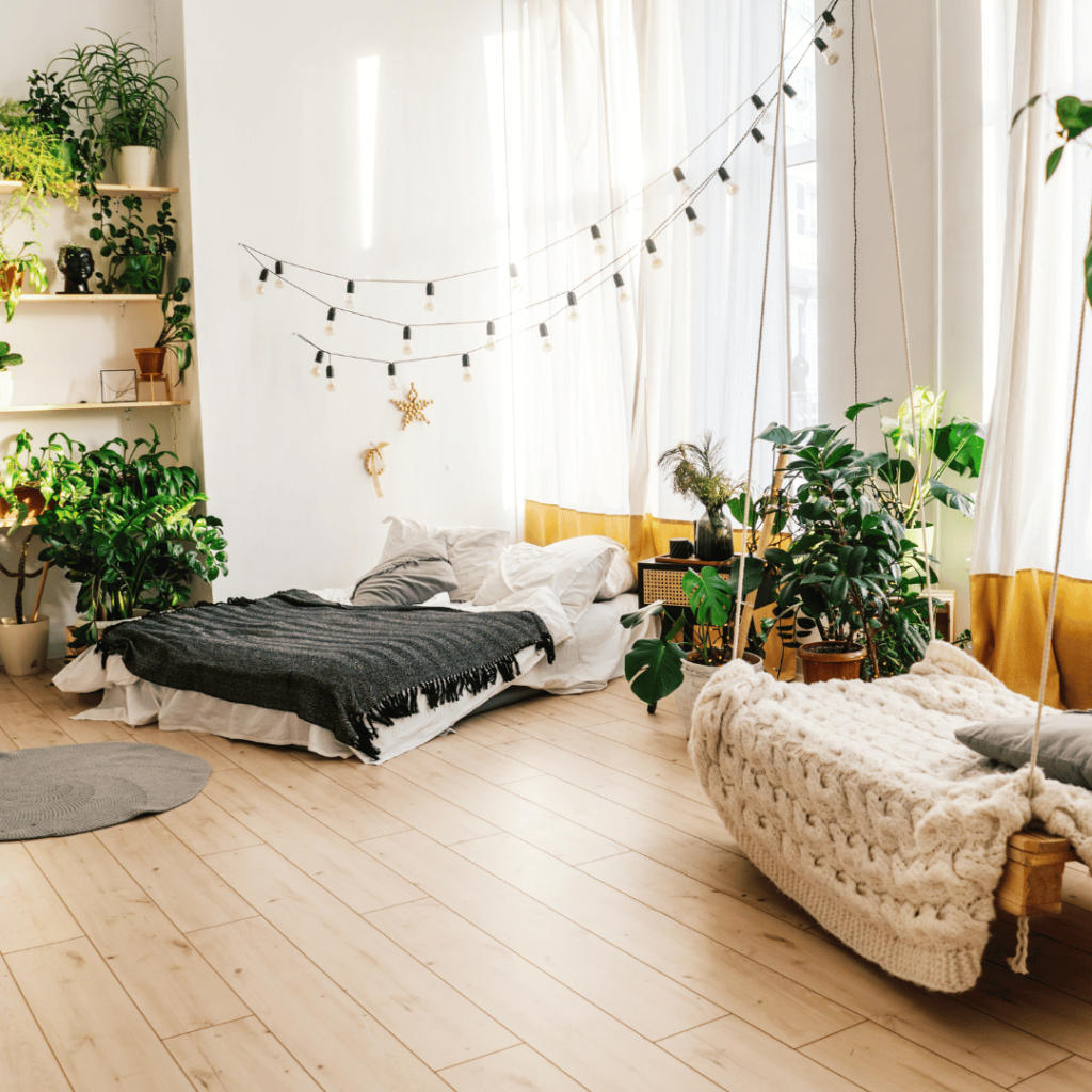 A bright and airy bedroom decorated in a bohemian style with biophilic elements. The room is flooded with natural light from sheer curtains and features a low-lying bed with crisp white bedding and a black throw. Plush knit blankets and earthy tones create a cozy atmosphere. An abundance of potted plants of various sizes and species brings life to the space, some resting on shelves and others on the floor. String lights are draped above, adding a whimsical touch to this peaceful, plant-filled sanctuary.