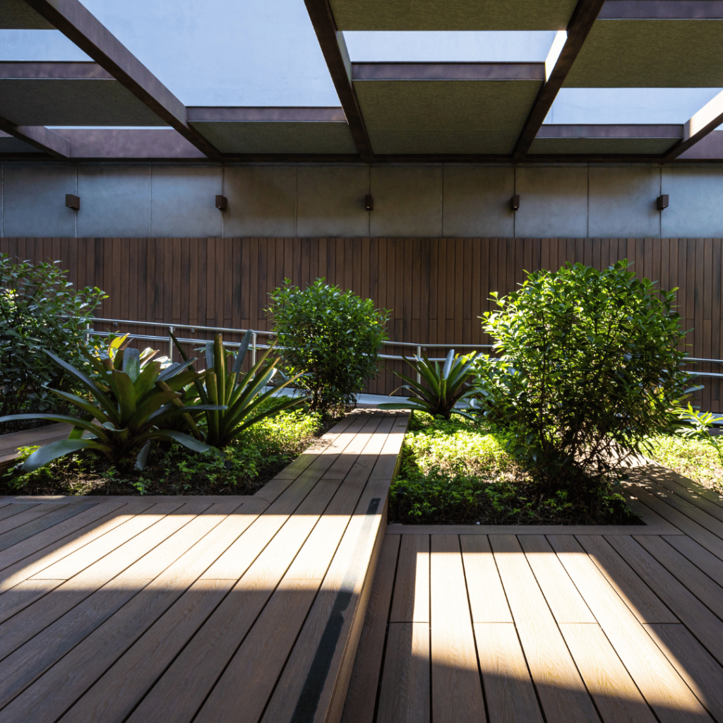 An urban garden path within a modern architectural setting, illuminated by natural light filtering through a pergola overhead. This intimate courtyard features wooden decking that guides the path through lush, green foliage and a variety of plants, including broad-leafed agaves. The garden is flanked by a dark wooden wall, creating a private, serene space that seamlessly blends the built environment with natural elements, epitomizing the essence of biophilic design.